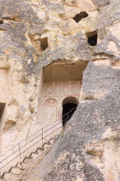 a yellow coloured rocky cliff face with a door and windows carved into it and some carvings and red-painted symbols around the door with wooden steps leading up to the door in goreme open air museum in cappadocia. historic places in turkey. 