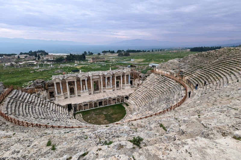 looking down from the top of srows of stone seats which are layed out in semi circular rows in Hieropolis Ancient Theatre in Turkey on an overcast day with light grey cloudy sky above.