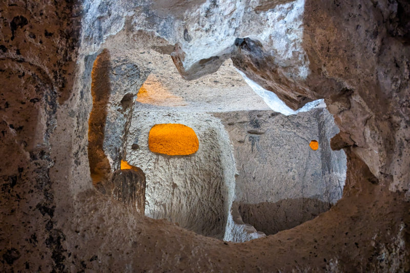 inside an underground cave with a rough circle cut out of a stone wall and a room on the other side of it where a small window cut into one of the the rocks has a light inside it. historic places in turkey. 