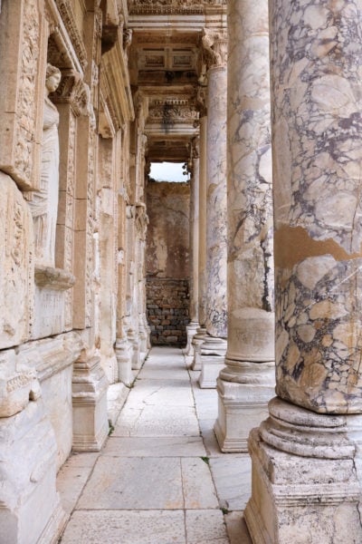 looking down a passageway between a beige stone wall with several white marble statues in alcoves along it on the left and a row of marble pillars along the right at the library of celsus in ephesus
