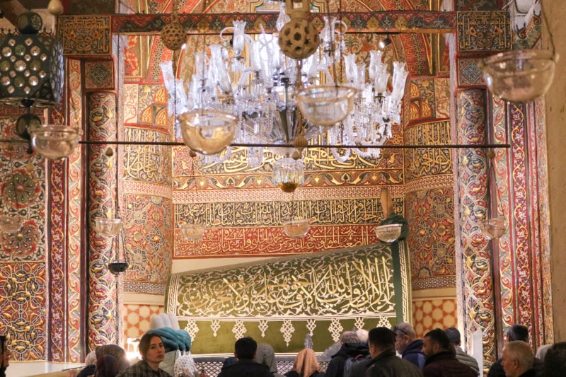 inside a mausoleum with walls covered in green orange and gold tiles and a large curved green tomb with gold lettering all over it and a glass chandelier above. there is a small crowd silhouetted in front of the tomb.
