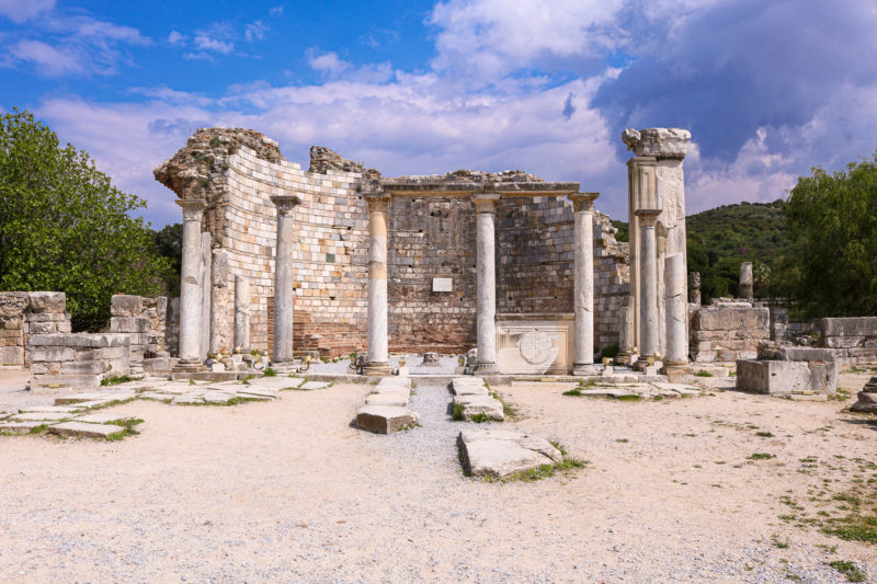 ruins of a curved brick wall with a cross carved into it and a row of 4 slim white stone pillars in front and the remains of a thicker white pillar to the right hand side on a sunny day with blue sky above. historic places in turkey to visit. 