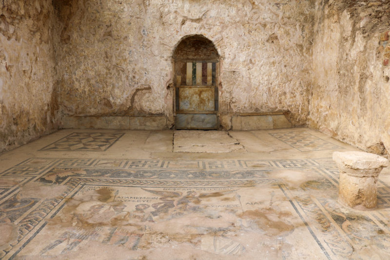 interior of an ancient room with bare beige stone walls and a large mosaic on the floor with mostly light brown tiles and a blue tiled design around the edge.