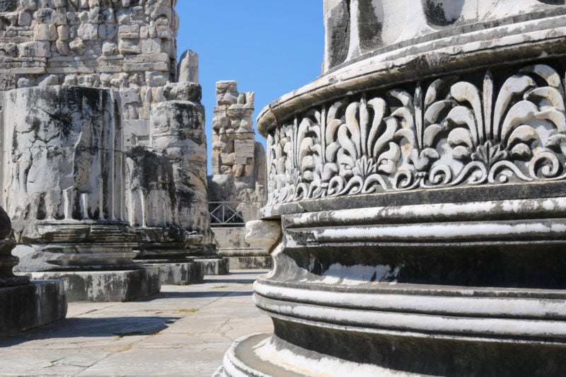 Close up of the base of a large white pillar with carvings around the base. there are more ruined pillars visible behind on a stone floor with a patch of blue sky visible. Historic places in Turkey. 