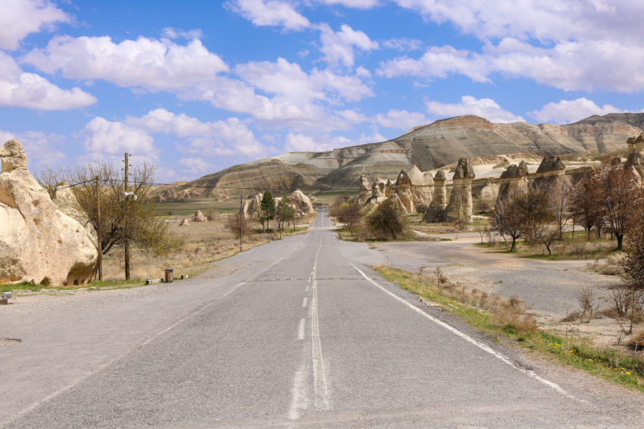 road leading through a dry desert-like landscape with conical shaed yellow and orange coloured rock stacks on either side of the road and a large brown hill in the distance on a sunny day with blue sky and fluffy clouds overhead. Near Zelve in Cappadocia Turkey.