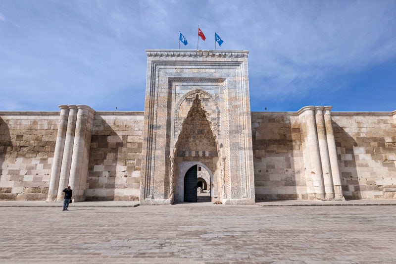 large cream marble building with a carved stone arch over the doorway and three flags flying from the roof against a blue sky - the caravanserai at sultanhan
