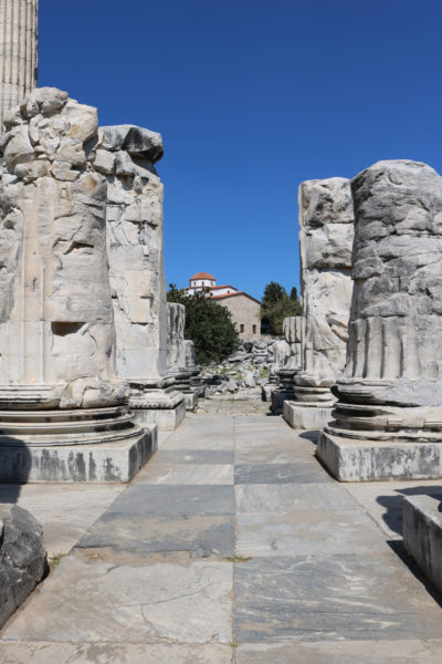 looking between two rows of half ruined white stone pillars with grey and white marble tiled floor between them and a small white building in the distance with terracotta roof with clear blue sky overhead