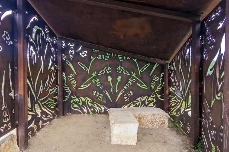 rusted metal wildlife blind with floral shapes cut out of the walls revealing greenery behind the blind