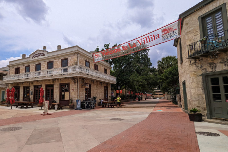 wide empty plaza with two historic stone buildings on either side of a walkway and a banner between them reading Welcome to la Villita in red text