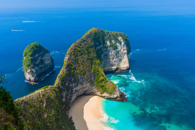 looking down from a cliff at a small headland topped with lush tropical greenery jutting out into a blue sea with a small white sandy beach sheltered behind the headland. Nusa Penida in Bali Indonesia.