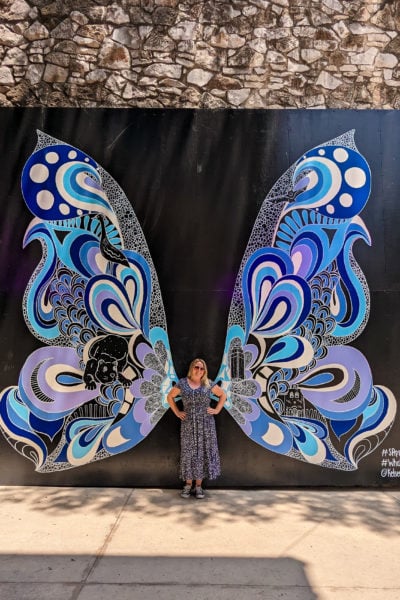emily wearing a long blue dress with small white flowers on it standing with her hands on her hips in front of a black wall with two large graffiti butterfly wings made up of different shades of blue - street art on the san antonio riverwalk