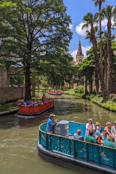 wide river in an urban area with two colourful open top boats filled with tour groups, the closest boat is turquoise and the further one is red, there is a church tower visible in the distance and lots of tall trees on either side of the river. The River Walk in downtown San Antonio