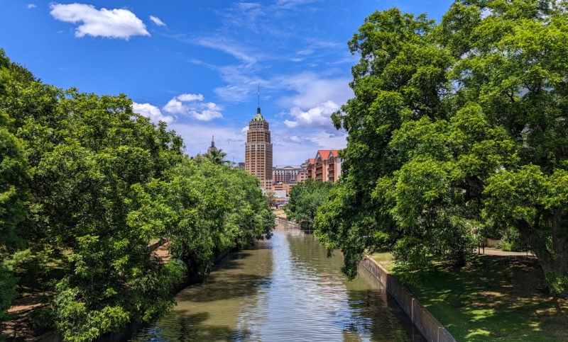 view of a wide river from a bridge with thick green trees on either side and part of a city skyline visible in the distance with blue sky overhead - day trip in San Antonio Texas
