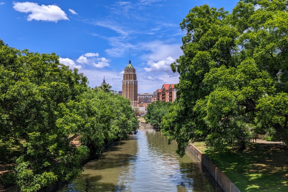 view of a wide river from a bridge with thick green trees on either side and part of a city skyline visible in the distance with blue sky overhead - reasons to visit San Antonio Texas