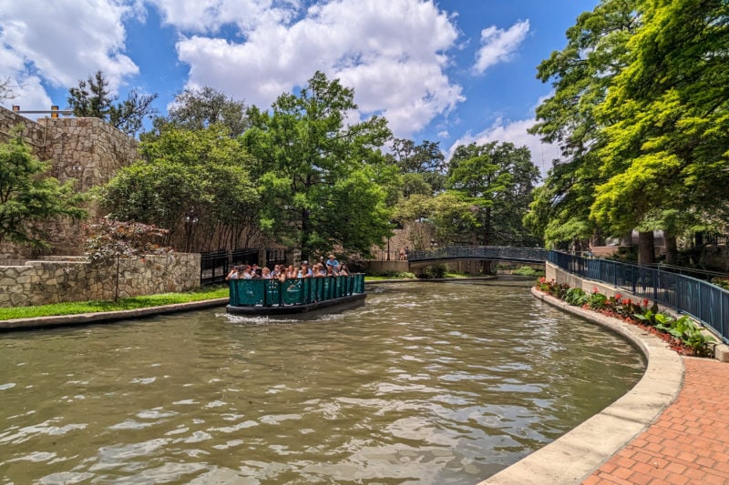 a wide dark green river with a paved walkway next to it an a small green open topped boat sailing down the river carrying a tour group. there are a lot of trees on either side of the river and blue sky overhead.