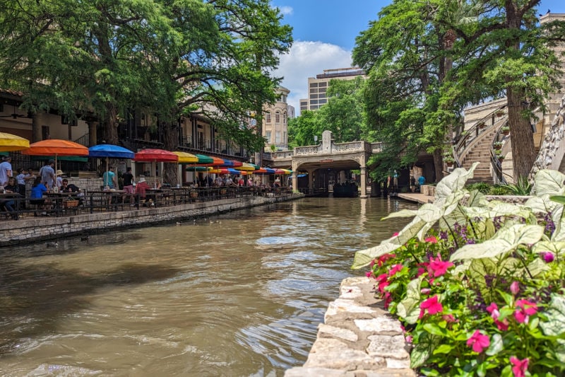 a wide dark green river with flowers on the bank closest to the camera and a row of tables with different coloured umbrellas on the opposite bank and a small stone bridge in the distance