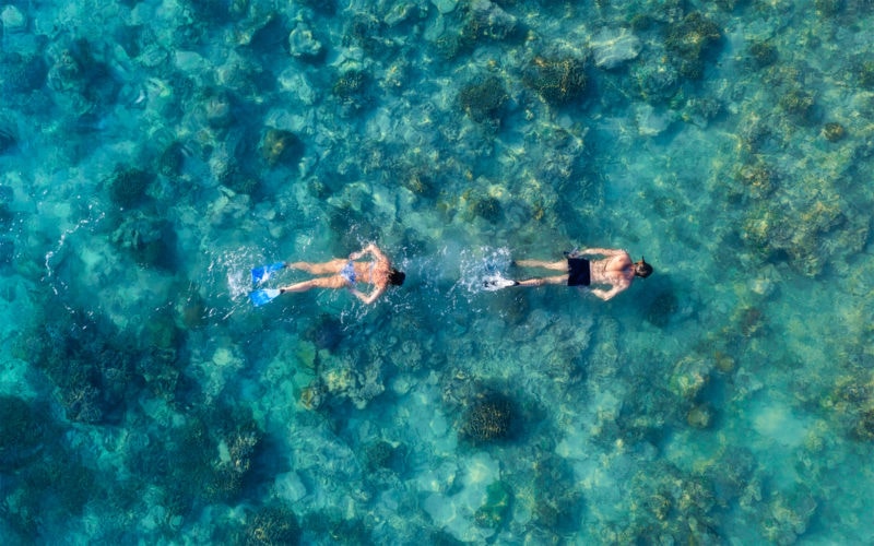 Aerial shot looking down at the sea where two men are snorkeling wearing blue flippers with very clear blue water below them dotted with coral - near Gili Meno Island. Best places to snorkel in Indonesia. 