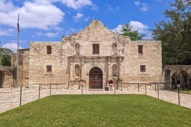 looking across a grassy lawn towards the Alamo in San Antonio, a very old church with beige stone walls and large wooden doors with blue sky and a few white clouds overhead
