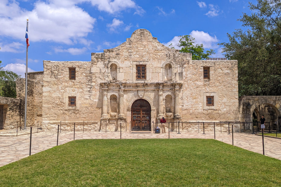 looking across a grassy lawn towards the Alamo in San Antonio, a very old church with beige stone walls and large wooden doors with blue sky and a few white clouds overhead