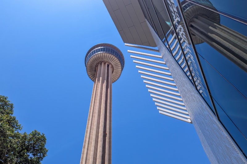 looking up at a very tall cylindrical tower with a circular building at the op against a bright blue sky - the tower is reflected in the windows of the all-glass building close to the camera