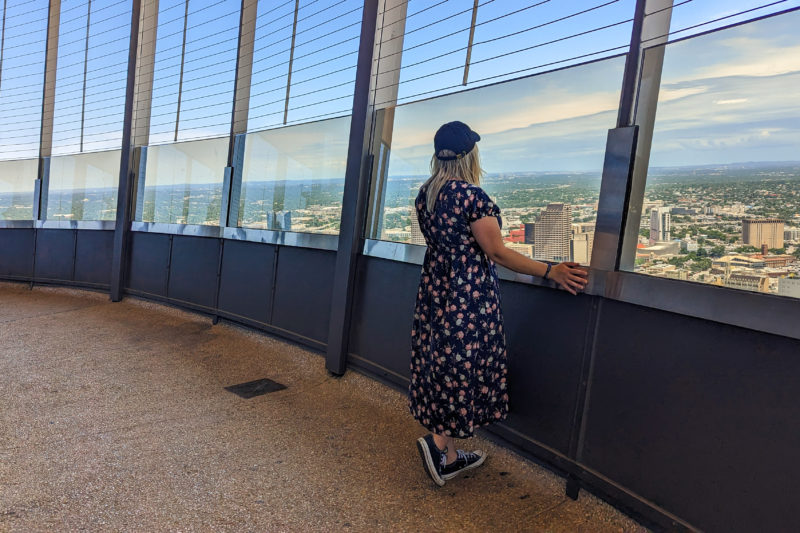 emily standing by a row of wide clouded windows at the top of a tall tower with a view of San Antonio city skyline below and blue sky overhead. emily is wearing a long navy blue dress with pink flowers and a black baseball cap and has her back to the camera with her long blonde hair down.