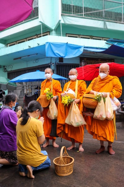 two thai women kneeling down at the edge of the street to pray after giving carrier bags filled with offerings to three thai bhuddist monks in bright orange robes. there are market stalls and a cyan coloured building behind them. 