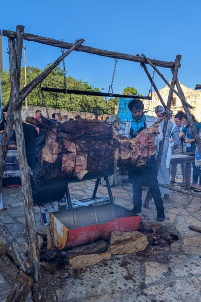 side of a cow hanging from a wooden frame above a fire pit in half of a red metal drum - cooking bbq outdoors in San Antonio on a sunny evening with blue sky above and a crowd of people behind