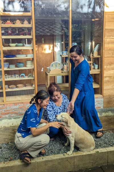 three thai women all wearing blue indigo dyed clothes in front of a small wooden shop, all three women are storking a golden retreiver dog, two are sat on the floor with the dog and the other is standing up