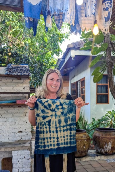 emily wearing a black dress and holding up a blue tie-dyed canvas bag in a small leafy courtyard with many blue dyed fabrics hanging up behind her. 