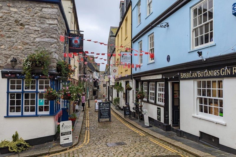 narrow cobbled street in Plymouth Barbican with a grey stone house on one side and a pastel blue painted shop on the other with red and white flag bunting overhead