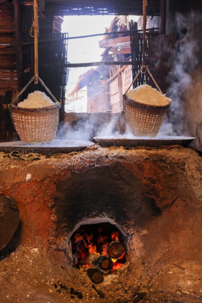 large wood burning oven built from earth or clay with two metal pans filled with white salty water on top and two woven baskets hanging above that with large piles of salt in each 