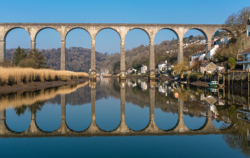 long stone viaduct across a still calm river perfectly reflected in the water with a small town on the bank on the right side and grassy fields on the left