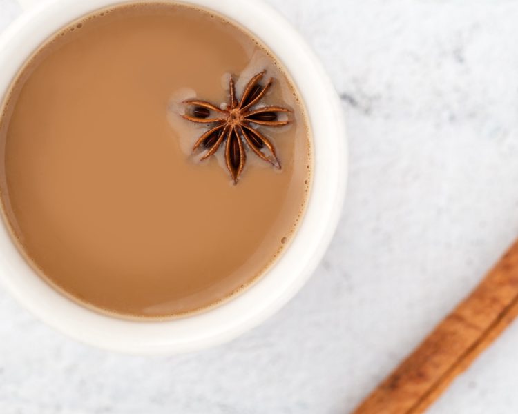 flatlay of a white mug with light brown chai tea and a single star anise floating in it on a white marble table next to a piece of cinnamon. Traditional Nepali drinks.