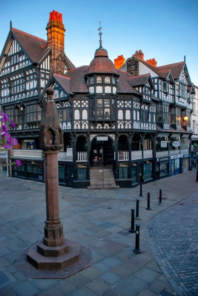 Chester Cross junction of grey flagstone streets with a large white and timber-clad Tudor building in the middle with three storeys and red tiled roofing. 