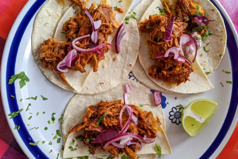 flat lay of three flour tacos on a white plate with a blue rim on a red table top. the tacos are topped with reddish-brown pork called cochinta pibil and pink onions with a wedge of lime next to them. 