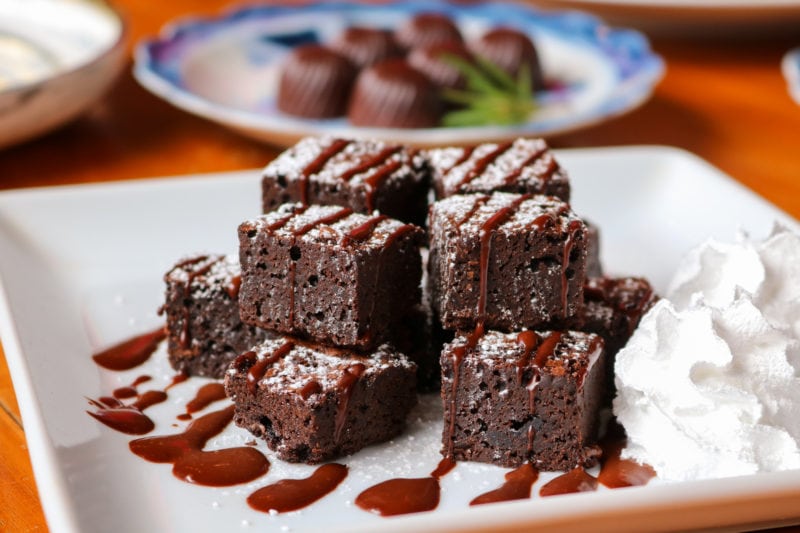 close up of a white square plate with a small pile of square brownies on it drizzled with chocolate sauce with a large blob of whipped cream next to them