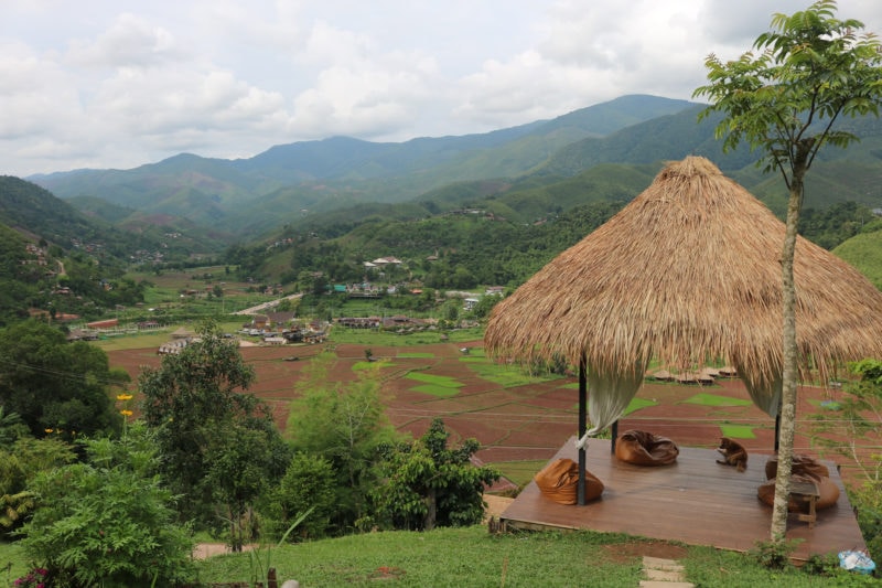 small wooden terrace with a grass roof above a few brown beanbags on the edge of a hill overlooking a small village surrounded by farmland and rice paddies with green mountains in the distance