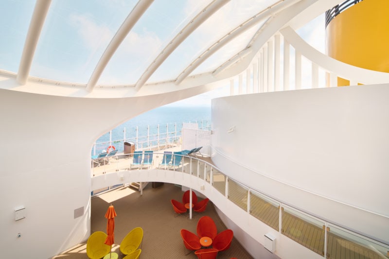interior deck of the costa esmerelda cruise ship with white curved walls and a glass panelled roof over a beige wooden deck with a few red and yellow tables on it