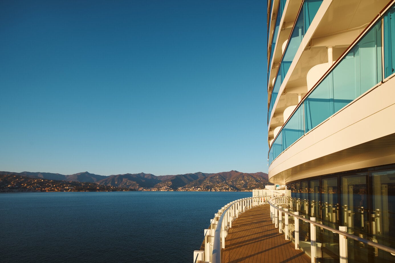 Exterior of the Costa Toscana cruise ship with a wraparound deck next to the side of the ship and the sea to the left with low green covered hills of the shore in the distance