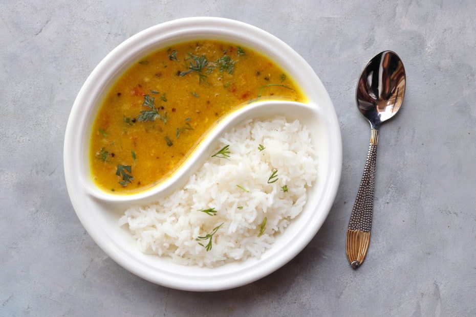 flat lay of a circular plate with two halves divided in a ying-yang shape. there is white rice in the lower half and a yellow-brown soup dal lentil in the top half with a metal spoon on the grey table next to the plate