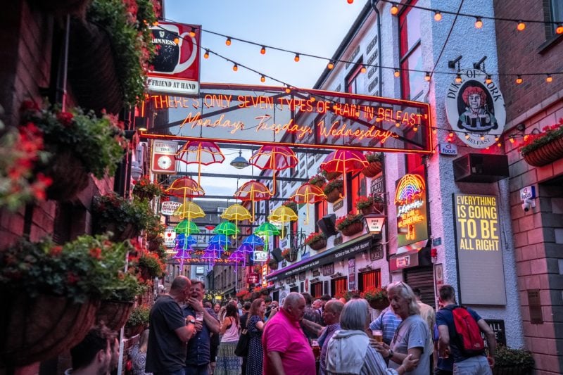 large crowd of people walking down a street in Belfast outside the historic white-washed Duke of York pub with strings of coloured lights over the street and a large neon sign that says there are only 7 types of rain in belfast, with rows of colourful lit up umbrellas behind. 