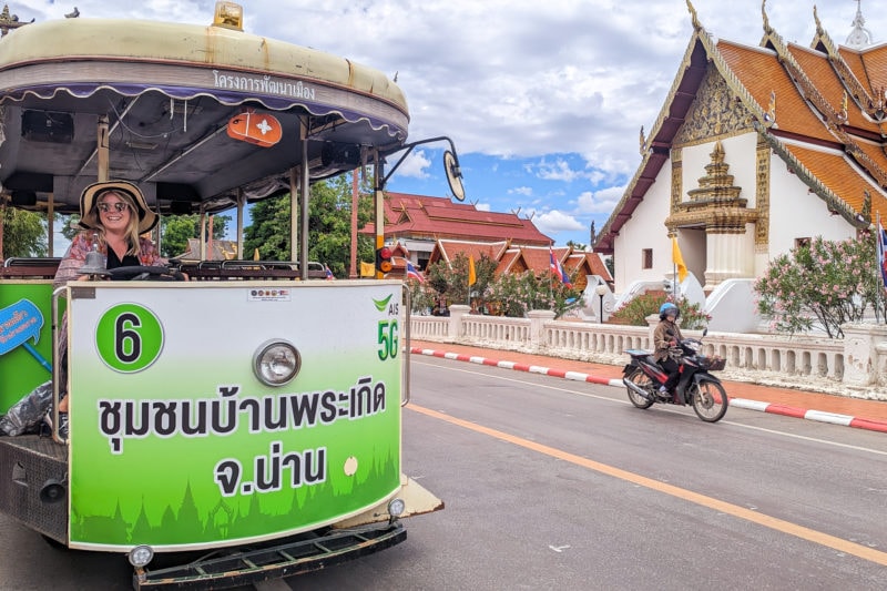 emily sitting in the drivers seat of a bright green painted electric tram on a road with a moped passing and a large cream temple behind with orange tiles triangle road. emily is wearing a floppy straw hat and an orange flowery shawl. the tram tour of nan town centre is one of the best things to do here. 