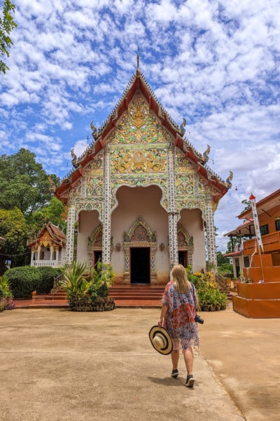 Emily wearing a long orange flowery shawl and holding a straw hat in one hand walking towards a small two storey temple with the exterior covered in green and gold gems and carvings against a blue sky
