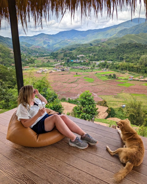 emily wearing denim shorts and a white short sleeved shirt sitting on a brown bean bag on a wooden terrace overlooking green farmland and paddyfields with mountains in the distance. there is a golden retriever sitting next to her on the wooden terrace and part of a grass frond roof overhead. In the village of Sapan in Nan Province Thailand. 