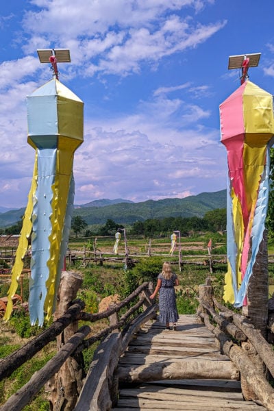 emily in a long navy blue dress with white flowers walking on a wooden bridge with green paddy fields beyond and mountains in the distance, there are two colourful silk decorations with long tails hanging up above the bridge in the foreground.