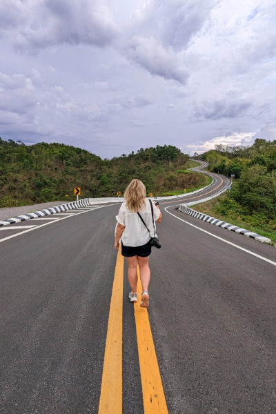 emily wearing denim shorts and a white short sleeved shirt walking away from the camera down the double yellow lines at the middle of a tarmac road which winds away over the hills into the distance. At Doi Phu Kha National Park in Nan Province Thailand. 
