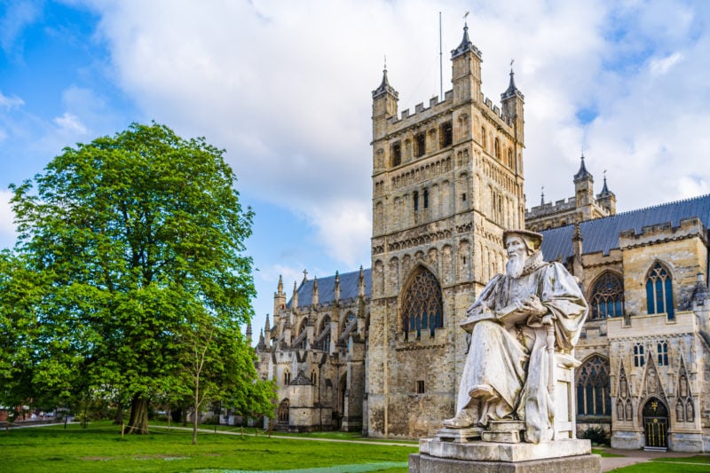 Grey stone statue of Richard Hooker in front of Exeter Cathedral, a large beige coloured stone building with a square shaped tower with four small spires on the top. there is a grassy lawn in front of the cathedral and a large leafy tree.