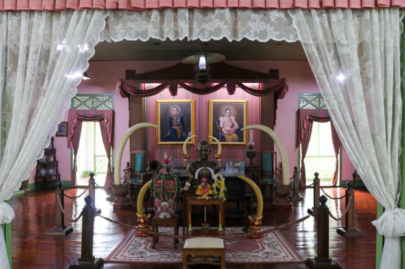 Interior of a bedroom in the Governor's Palace in Phrae Thailand with pink painted walls and a large pink four poster bed with lace trim. there are paintings on the wall and dark pink curtains around the windows