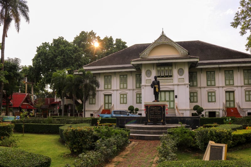 Exterior of the Governor's Palace in Phrae Thailand - a large two storey white mansion built from wood with pale green trim and a grey tiled roof. the sun is setting behind a tree next to the house with lens flare coming through the branches. there is a large lawn and hedges in front of the house.