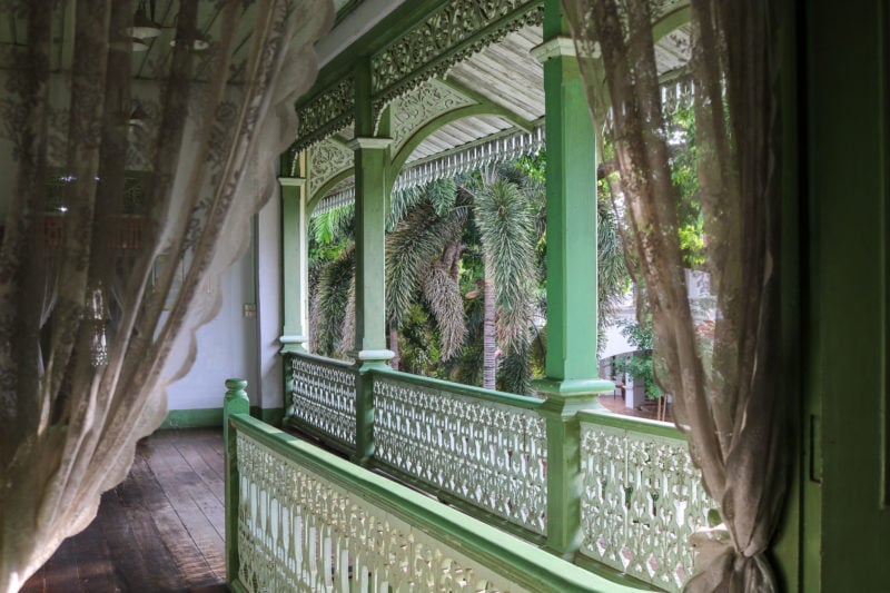 upper floor balcony at the Phrae Governor's Palace with green painted wooden pillars and a white painted wooden railing. there are lace curtains in the foreground of the shot and palm tree leaves visible behind the balcony.  things to do in phrae thailand.